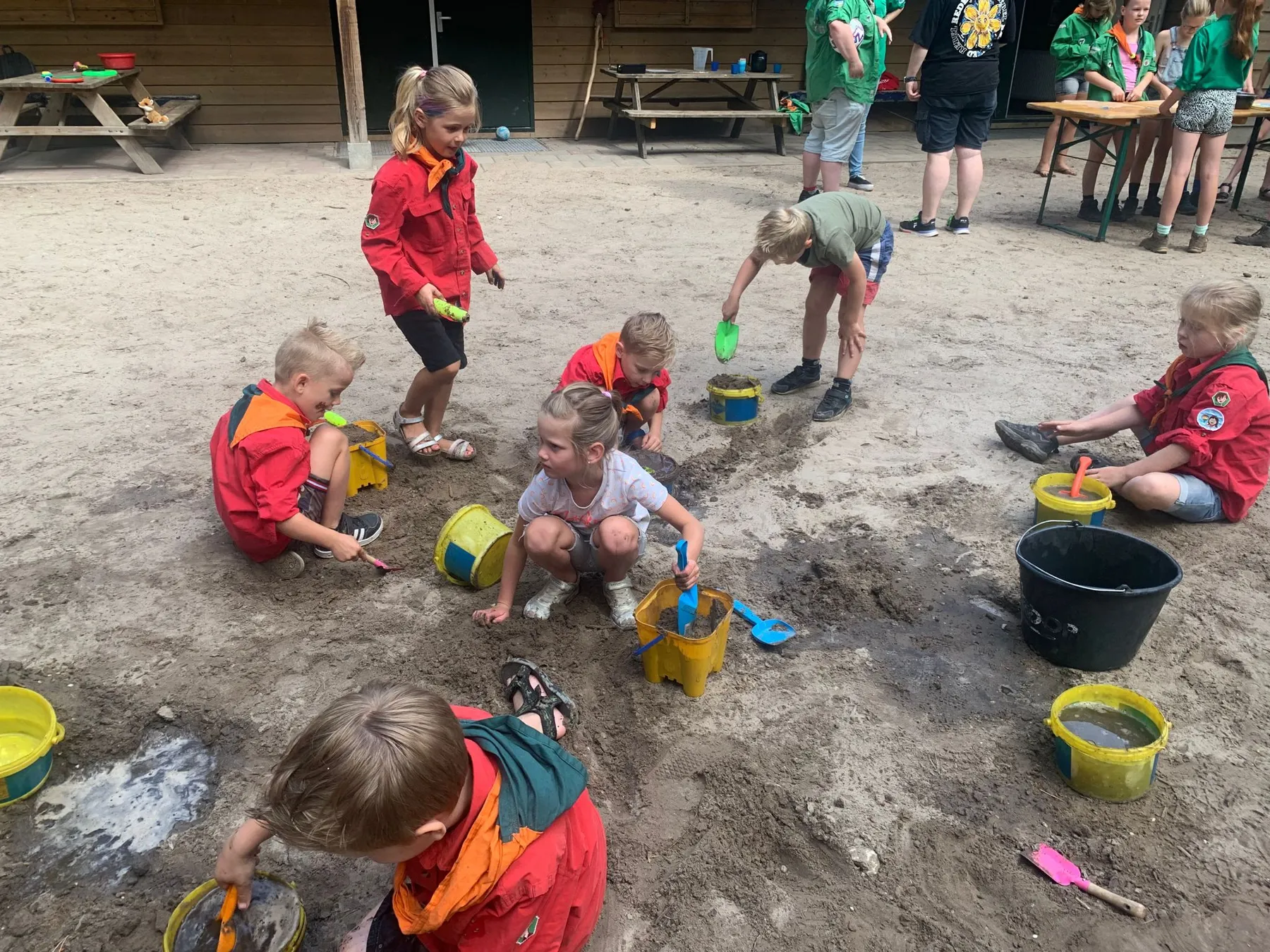 Bevers in rode blouses spelen met emmers en schepjes in het zand voor het clubhuis
