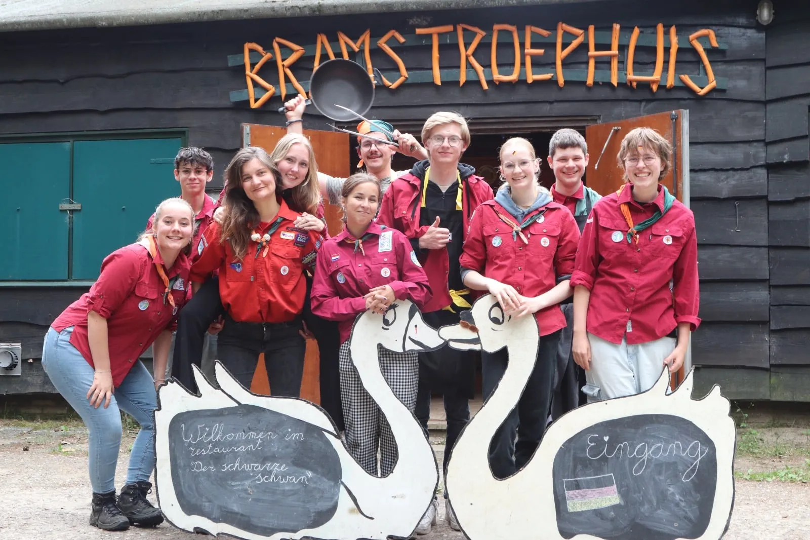 Lachende jongeren in rode scoutingblouses voor Brams Troephuis, achter twee houten zwanen met krijtborden