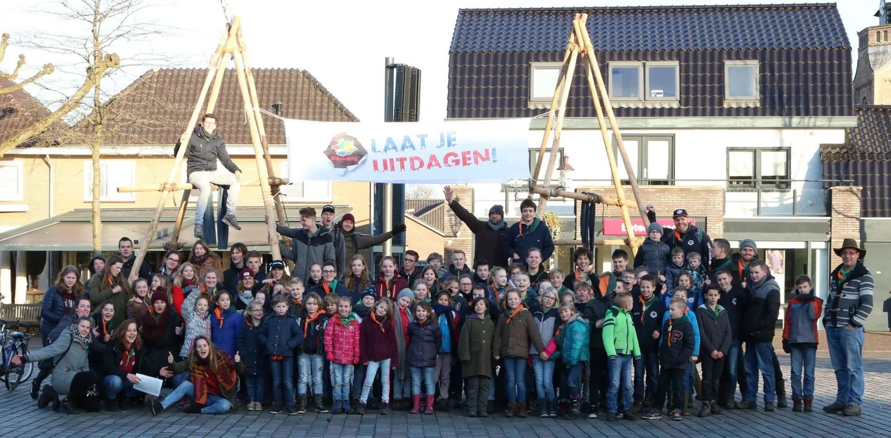 Grote groep scouts van alle leeftijden op een plein in Nunspeet, onder een spandoek met 'Laat je uitdagen!' tussen twee zelfgebouwde houten torens