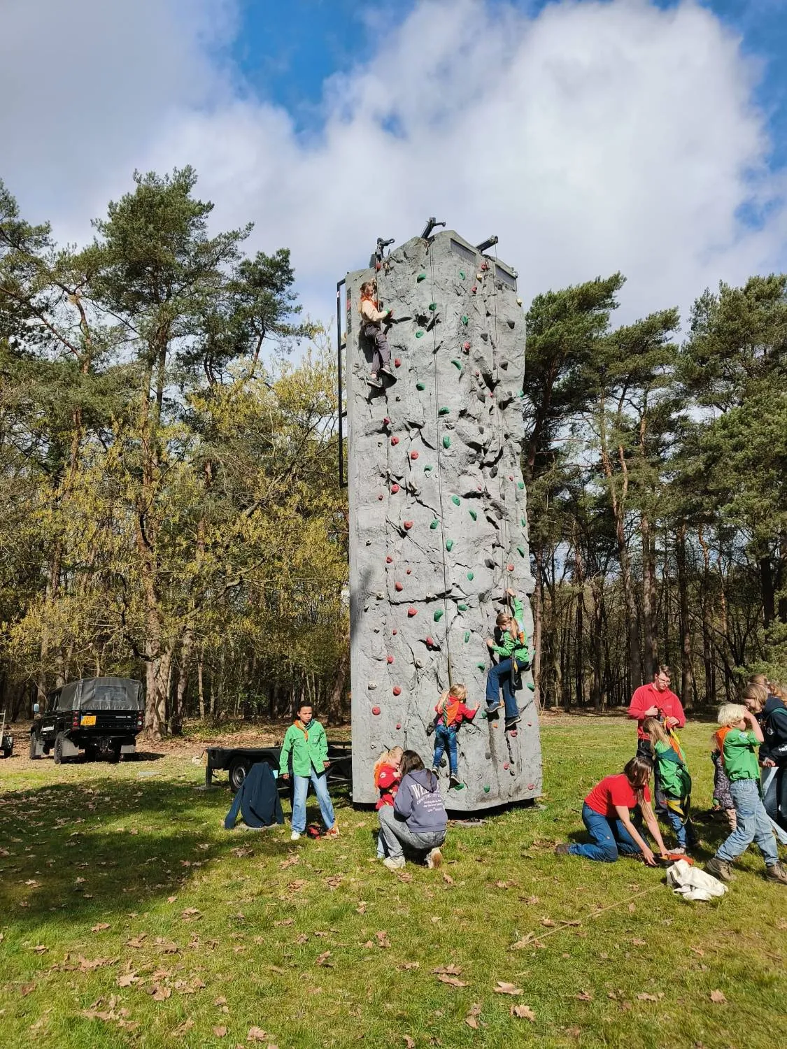 Welpen in groene blouses klimmen aan touwen tegen een hoge klimwand in het bos