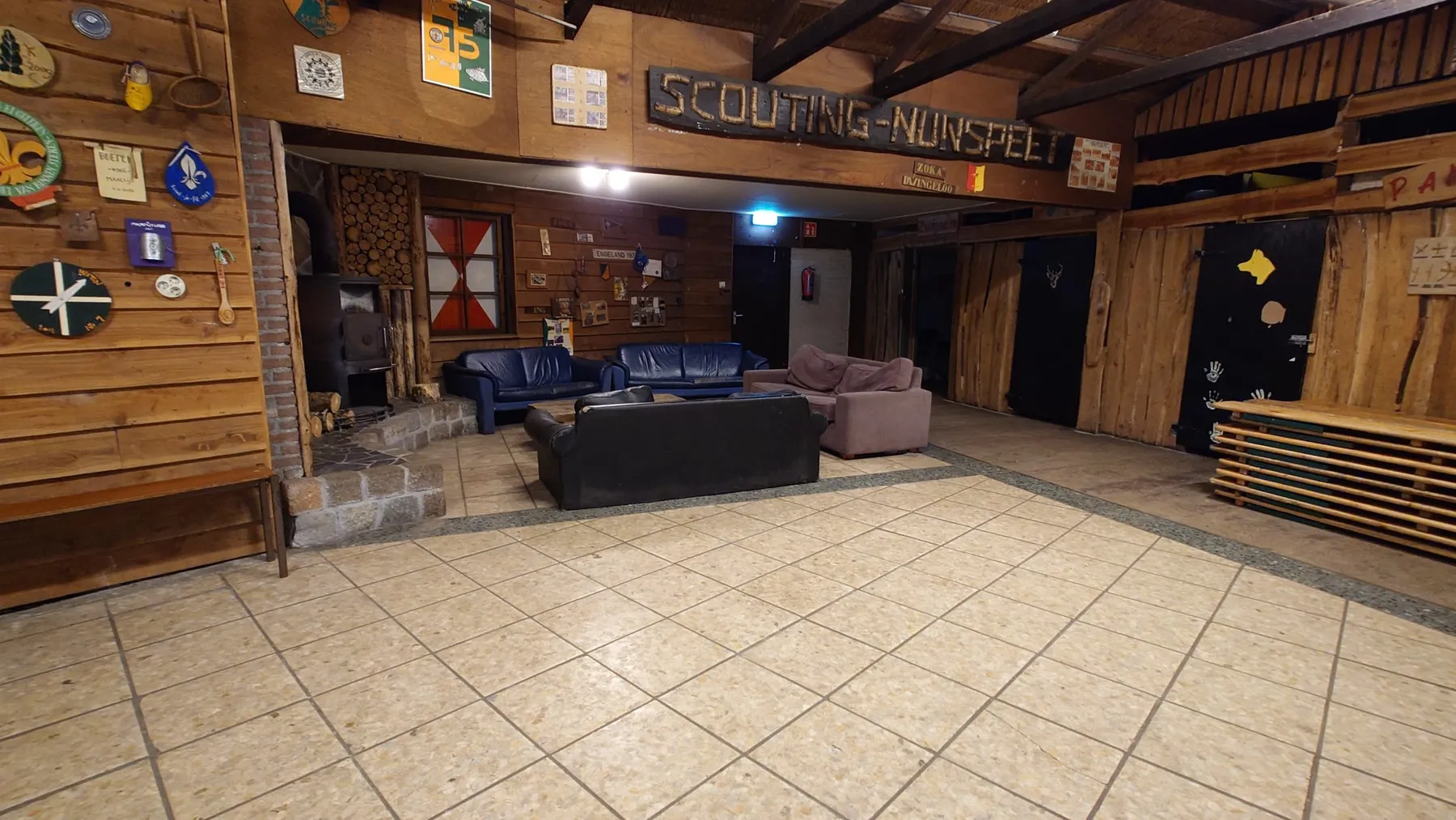 Grote zaal met houten wanden, bord Scouting Nunspeet, houtkachel en een zithoek met banken